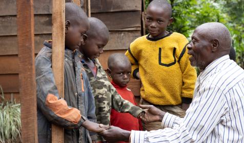 Michel sits outside holding hands with his grandchildren, who stand beside him.
