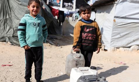 Two young boys standing with water canisters. In the background is the temporary shelters for displaced people in Gaza
