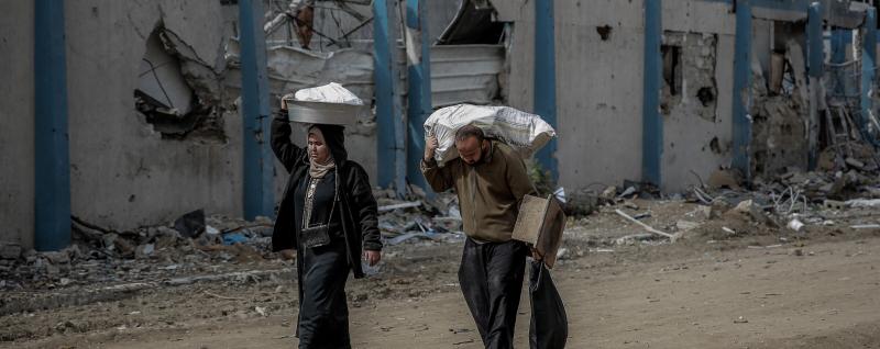 Man and woman carry supplies walking past buildings destroyed by missile strikes. Sina square, Gaza City, 21 February 2024