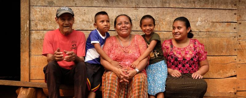 Aurelia and her family sit on a wooden bench outside their home, smiling. She holds a young boy while a little girl hugs her. A man in a red t-shirt and a woman in a pink blouse smile beside them.