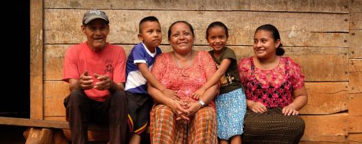 Aurelia and her family sit on a wooden bench outside their home, smiling. She holds a young boy while a little girl hugs her. A man in a red t-shirt and a woman in a pink blouse smile beside them.
